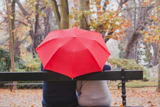 Two people sitting closely together on a park bench, sharing a bright red umbrella representing umbrella insurance coverage.