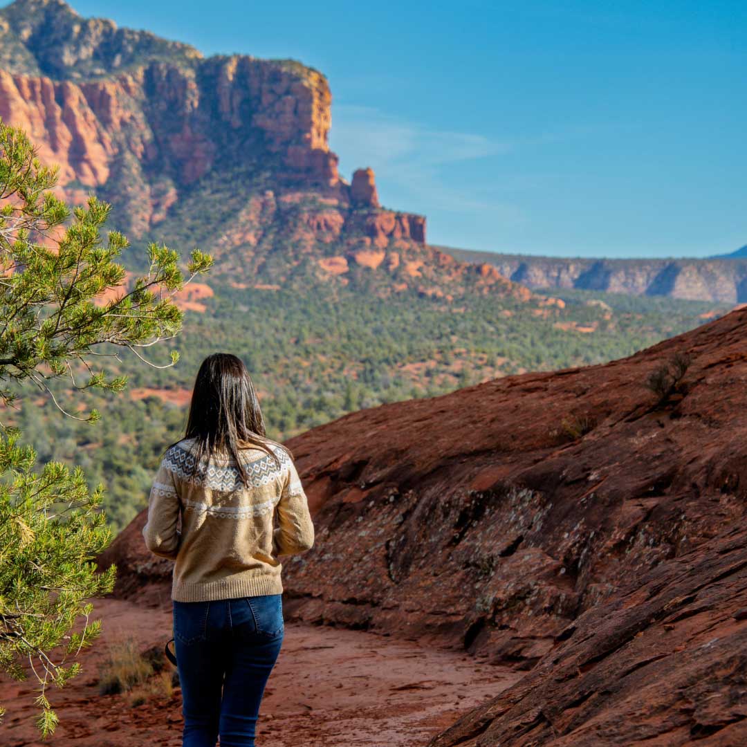 A person with long dark hair wearing a patterned beige sweater and blue jeans stands on a red rocky trail illustrating personal insurance protection.