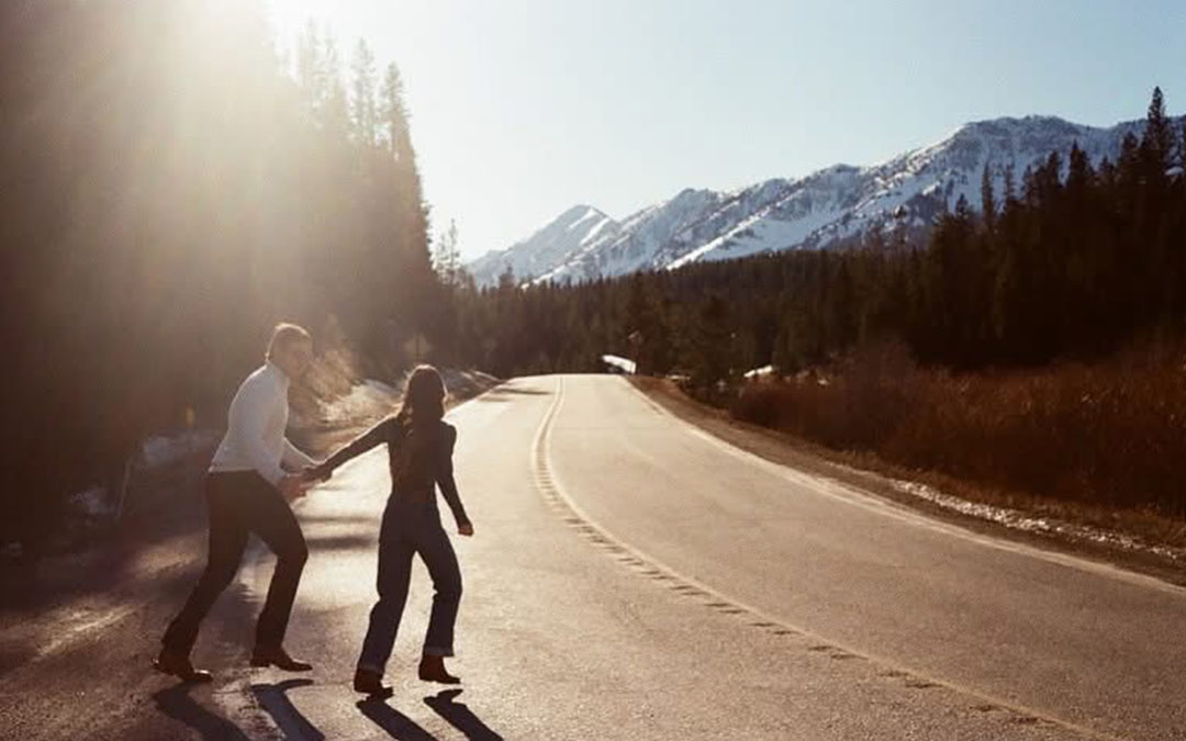 A couple strolls down a road, with majestic mountains rising in the background under a clear blue sky