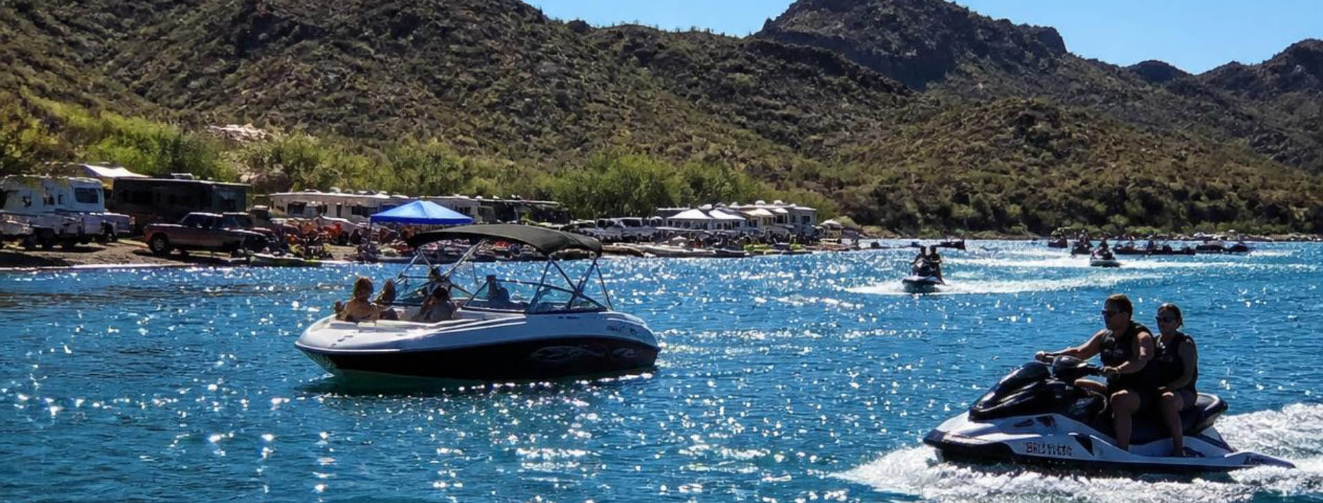A motorboat and jet skis navigating a sunny, blue Arizona lake with a crowded shoreline of campers and RVs under desert mountains.