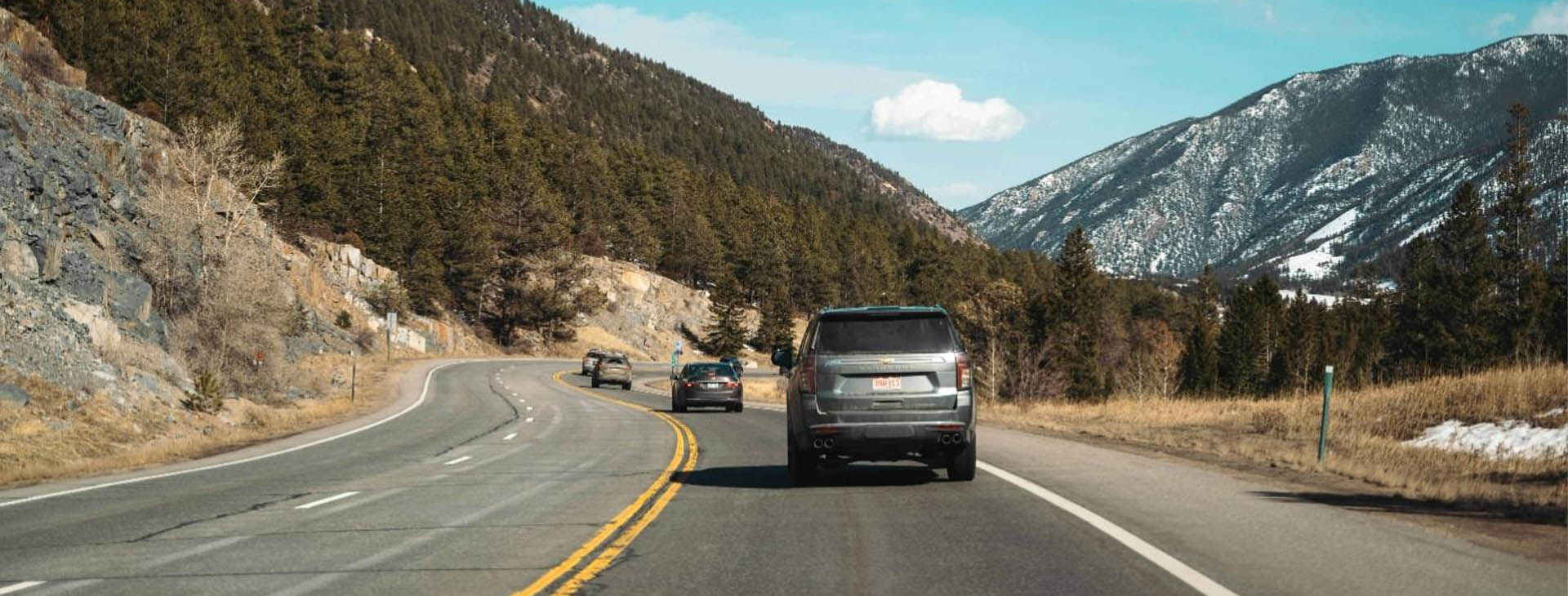Vehicles traveling along a scenic mountain highway surrounded by pine trees and snow-capped peaks, representing Montana vehicle insurance coverage.