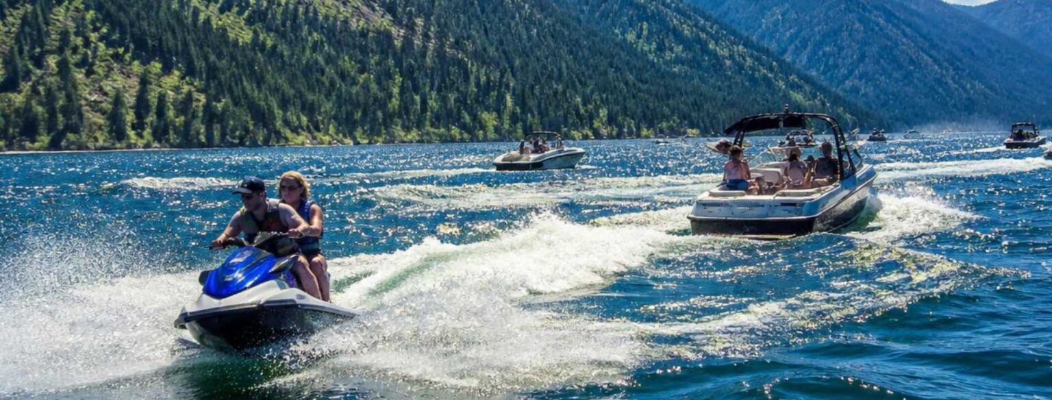 Two people riding a blue jet ski and a motorboat filled with passengers cruising on a large Montana lake surrounded by dense evergreen pine forests.